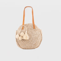 Round raffia handbag with pom-pom decoration and adjustable leather shoulder straps. Fully lined with inside pocket. Available in beige.