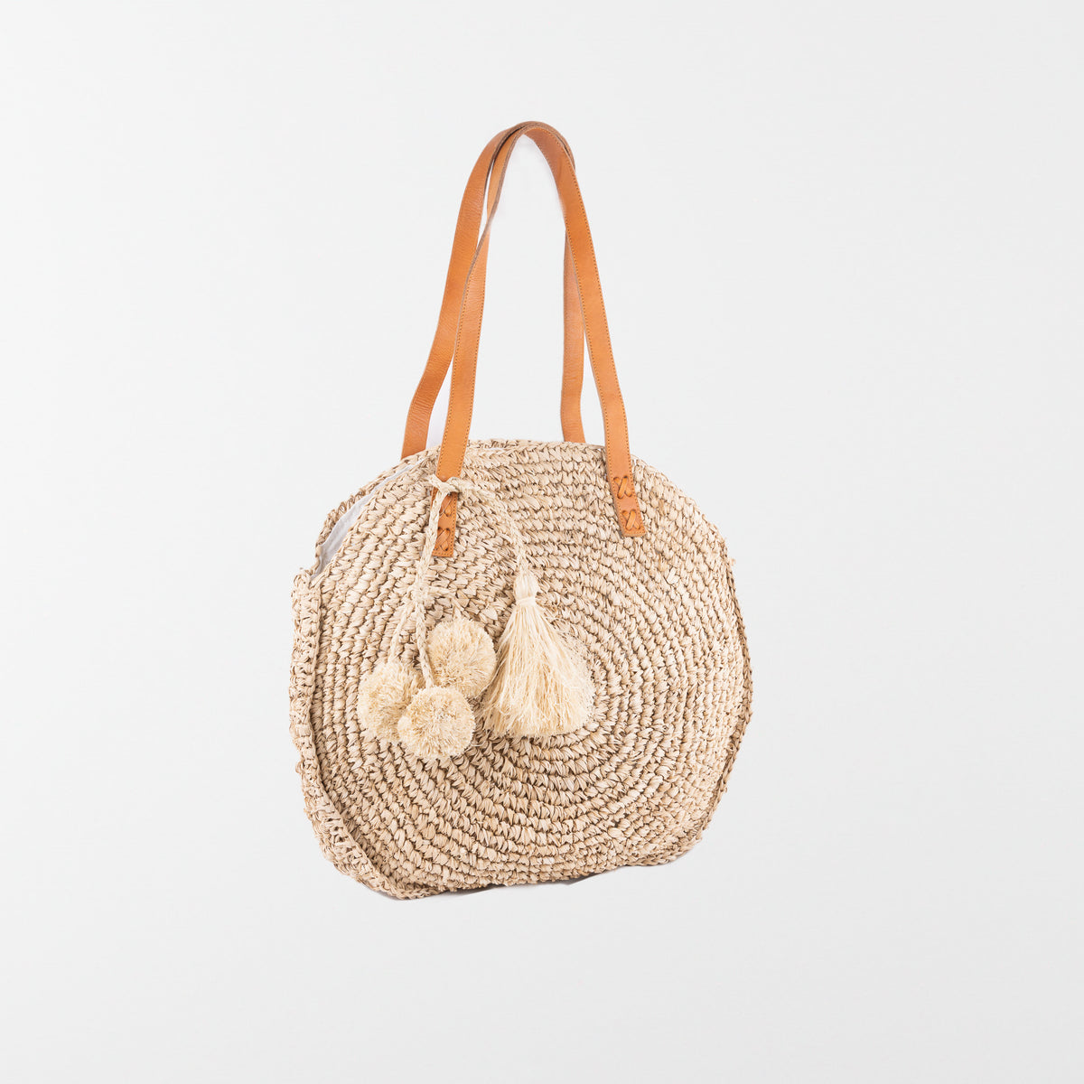 Round raffia handbag with pom-pom decoration and adjustable leather shoulder straps. Fully lined with inside pocket. Available in beige.